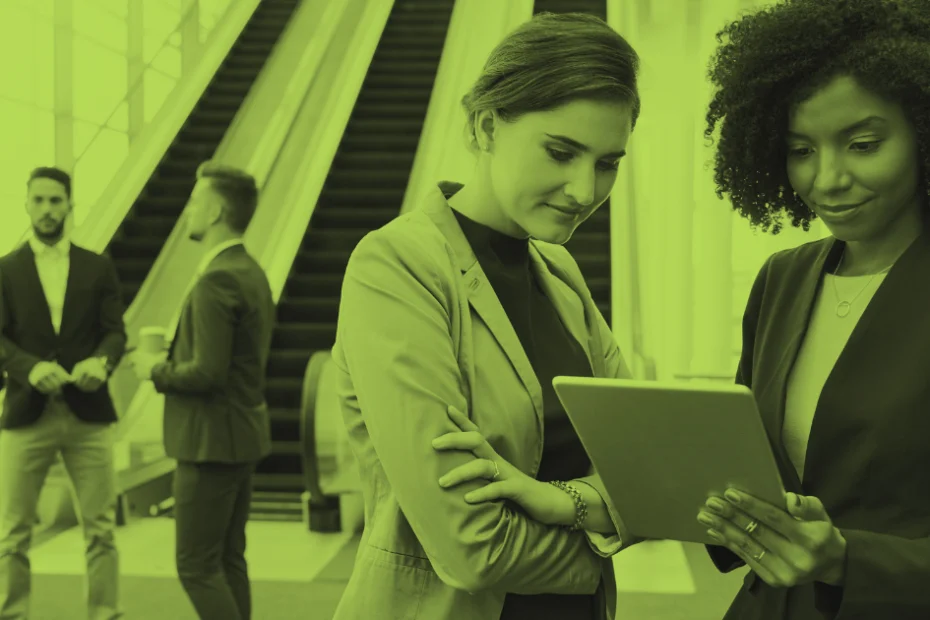 Two businesswomen reviewing information on a tablet in a modern office lobby, with other professionals conversing in the background.