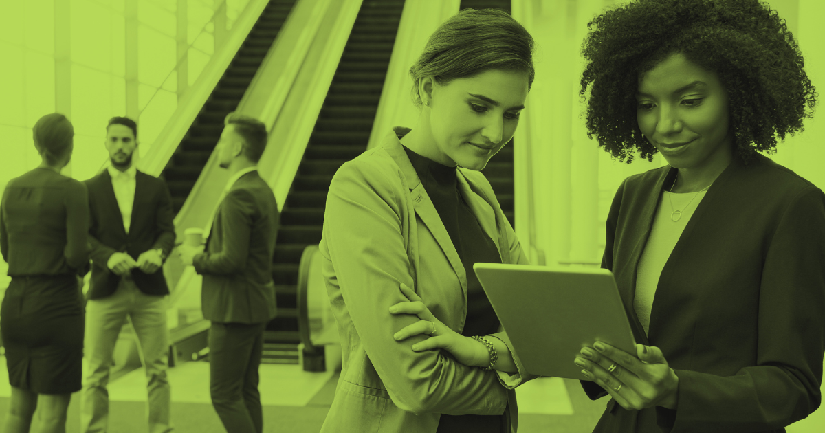 Two businesswomen reviewing information on a tablet in a modern office lobby, with other professionals conversing in the background.