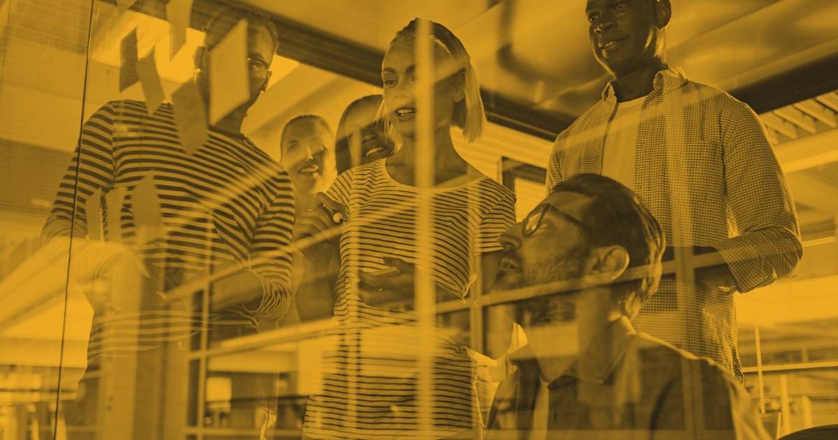 A group of colleagues gathered around a glass wall, collaborating and discussing ideas in an office setting.